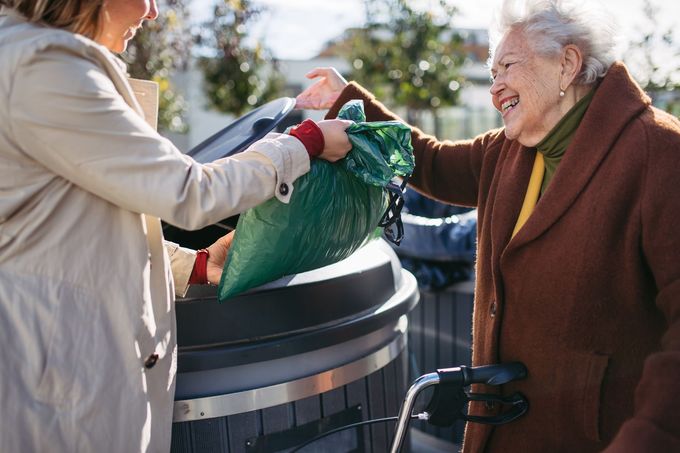 Two people place a green trash bag into a large outdoor bin on a sunny day.