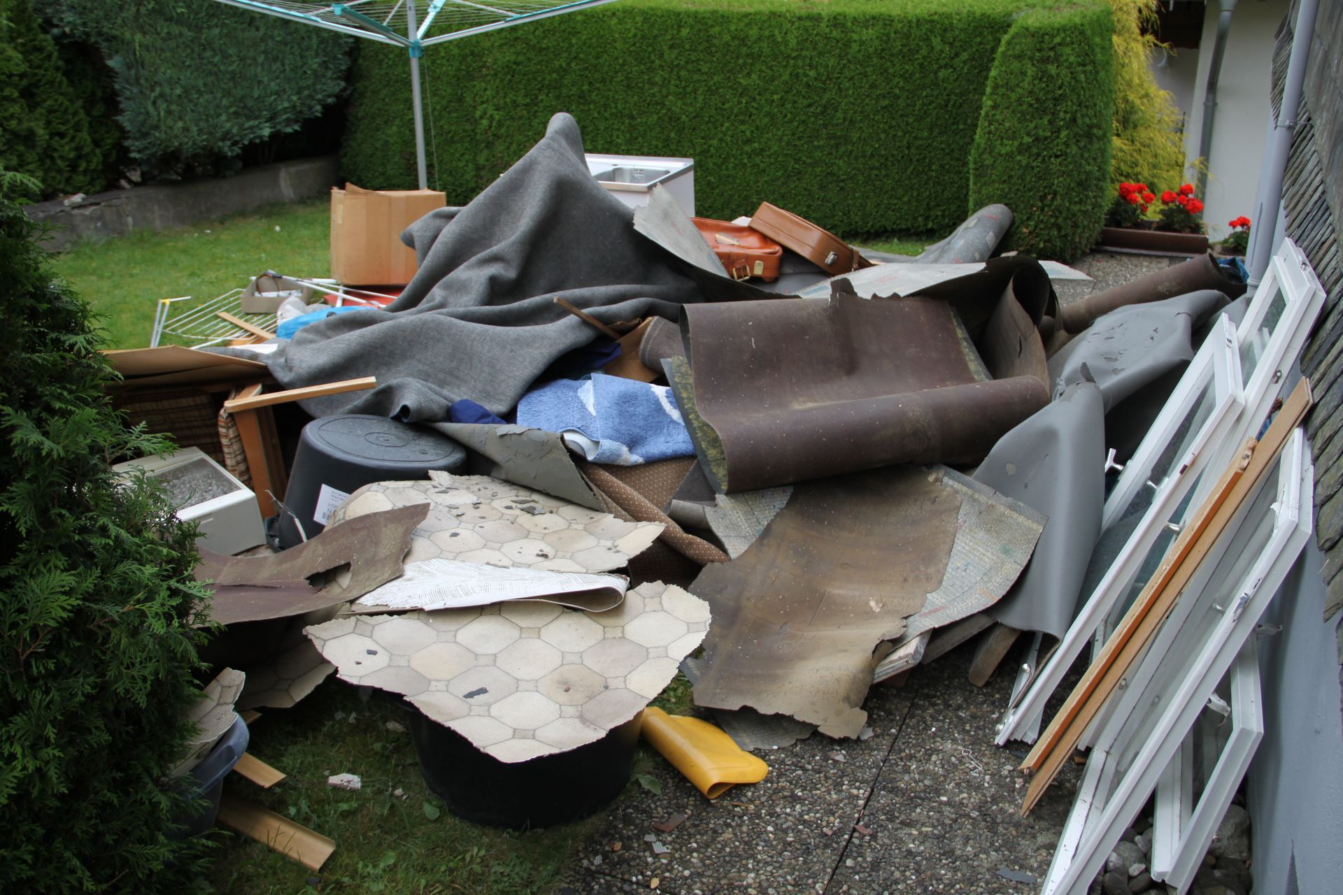 Pile of construction debris and rolled roofing material beside a hedge and fence outdoors