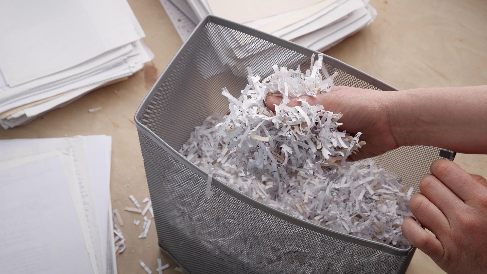 Hands shredding white paper into a gray mesh bin beside stacks of paper