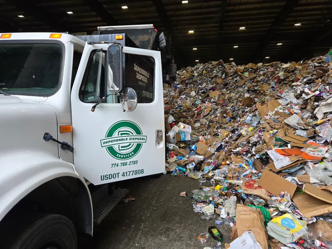 White garbage truck in a landfill beside a huge pile of mixed trash inside a warehouse-like facility
