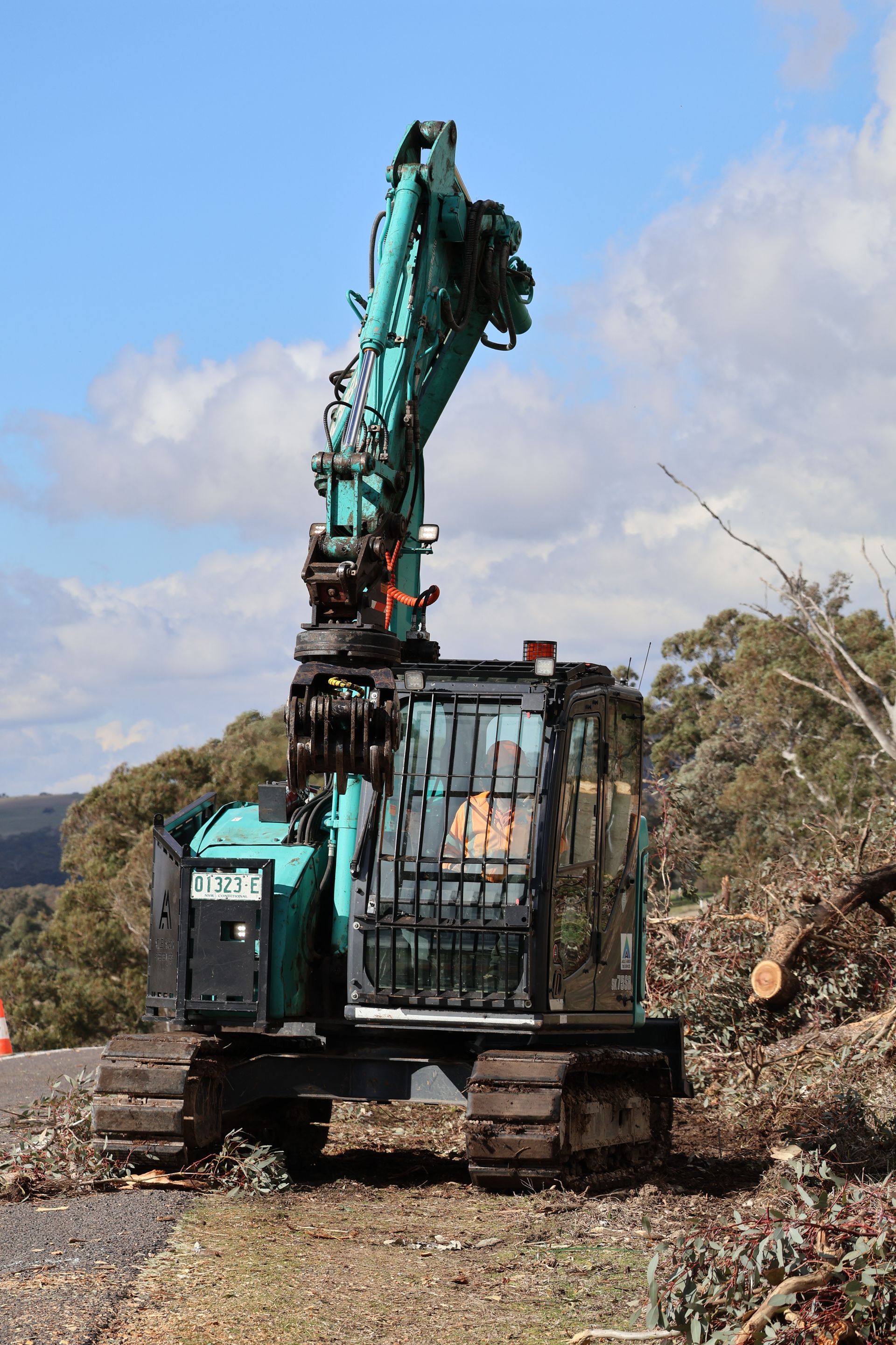 A teal forestry processor with tracks and a protected cab sitting on dirt near trees under a blue, cloudy sky.