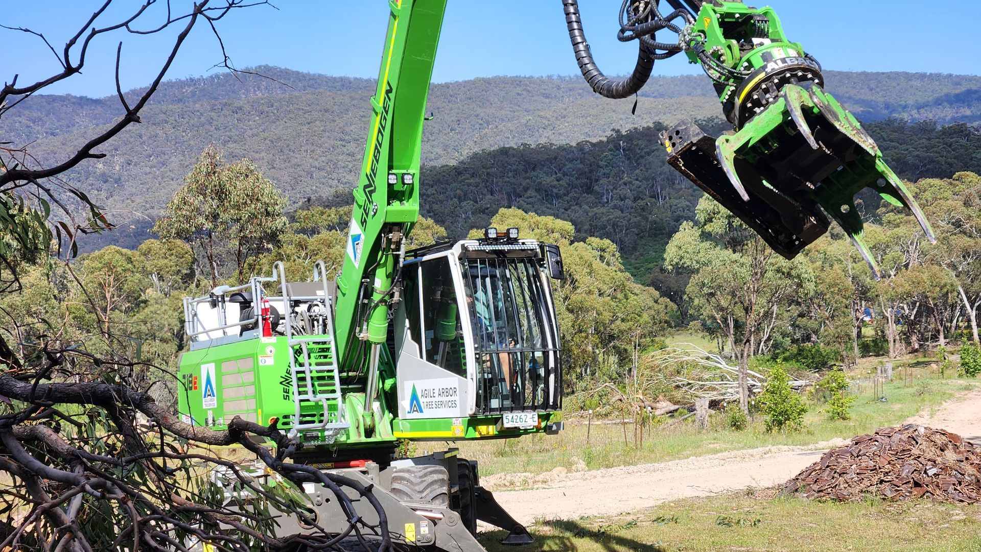 A bright green Sennebogen material handler with a grapple attachment operates in a wooded, mountainous landscape.