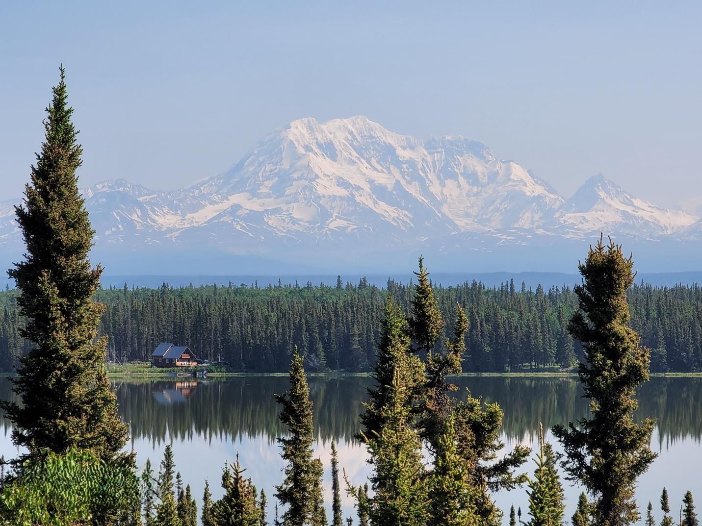 Snow-capped mountain range rises above a forest and lake, with a cabin visible.