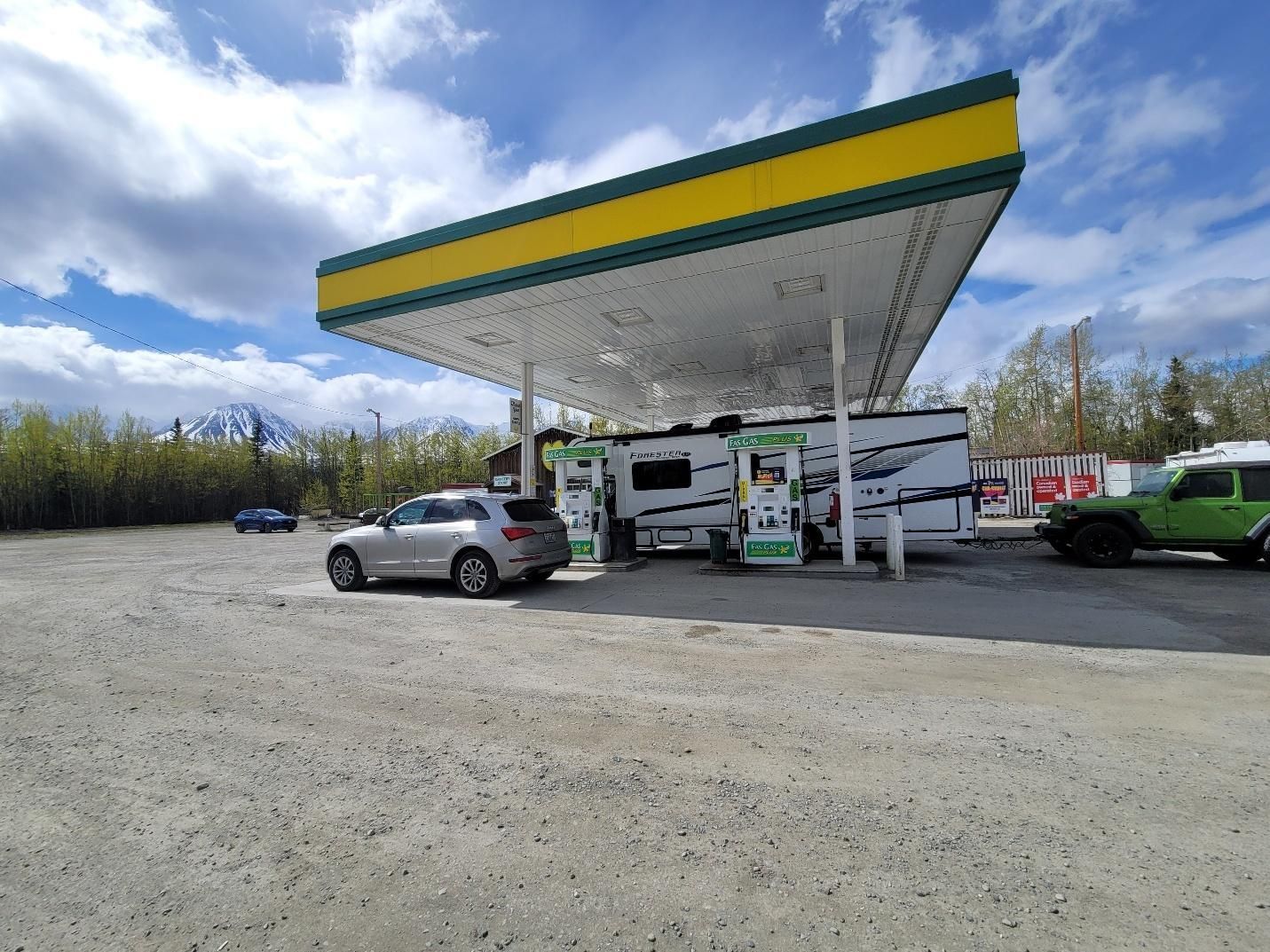 A car and RV at a gas station with a yellow and green canopy, under a cloudy sky.