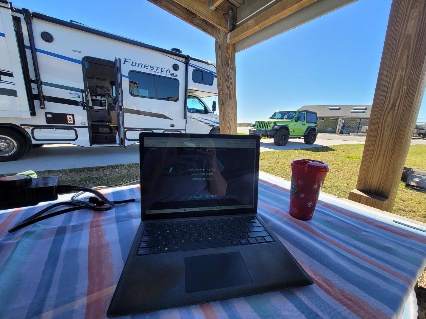 Laptop on a striped table, RV and green Jeep in the background on a sunny day.