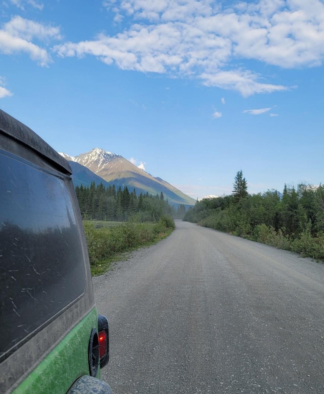 A green vehicle drives down a gravel road toward a mountain under a blue sky.