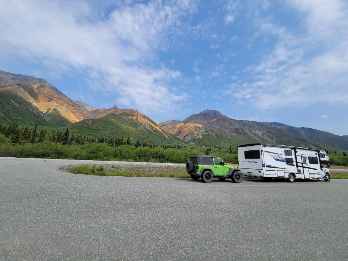 Green Jeep and white RV parked in gravel lot with mountains and blue sky.