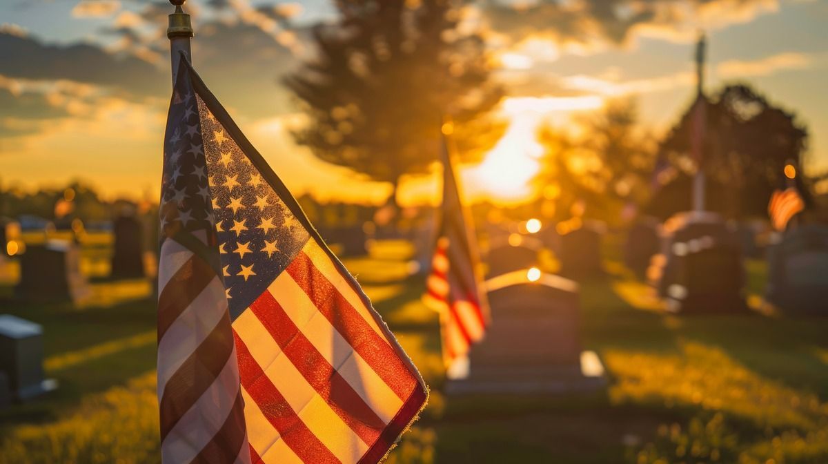 American flags stand before headstones in a cemetery at sunset, casting a warm, golden glow over the scene.