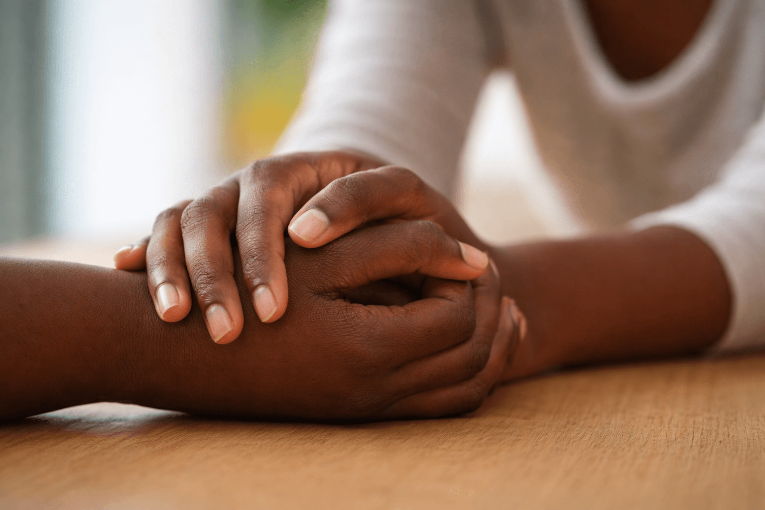 Two hands gently hold one another over a wooden surface, symbolizing comfort and support.