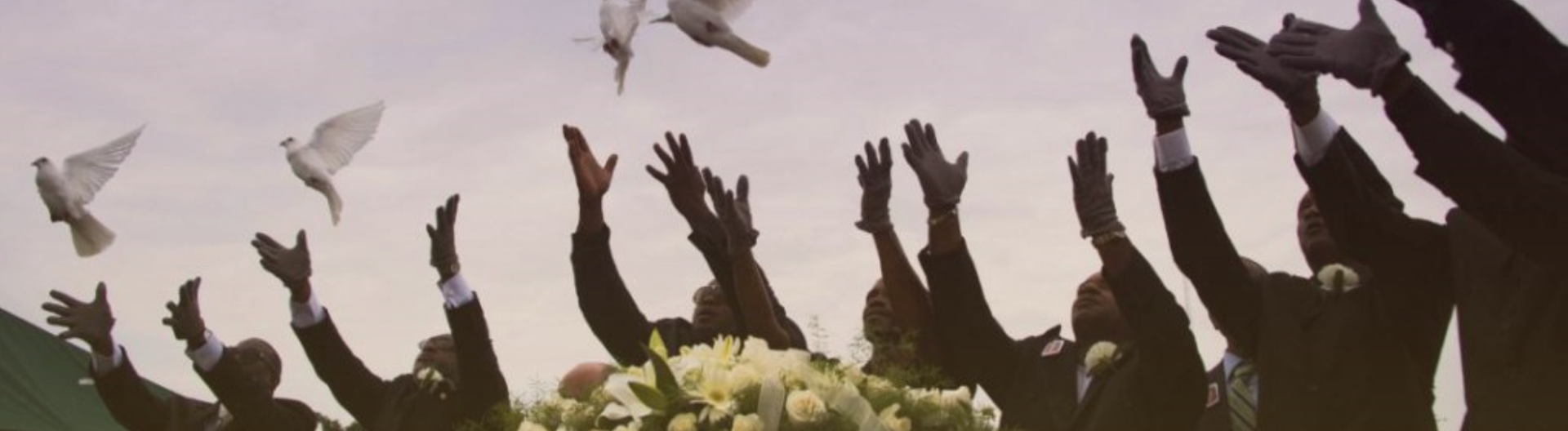 A group of people in suits release white doves into the sky, their arms raised in a gesture of celebration or tribute.