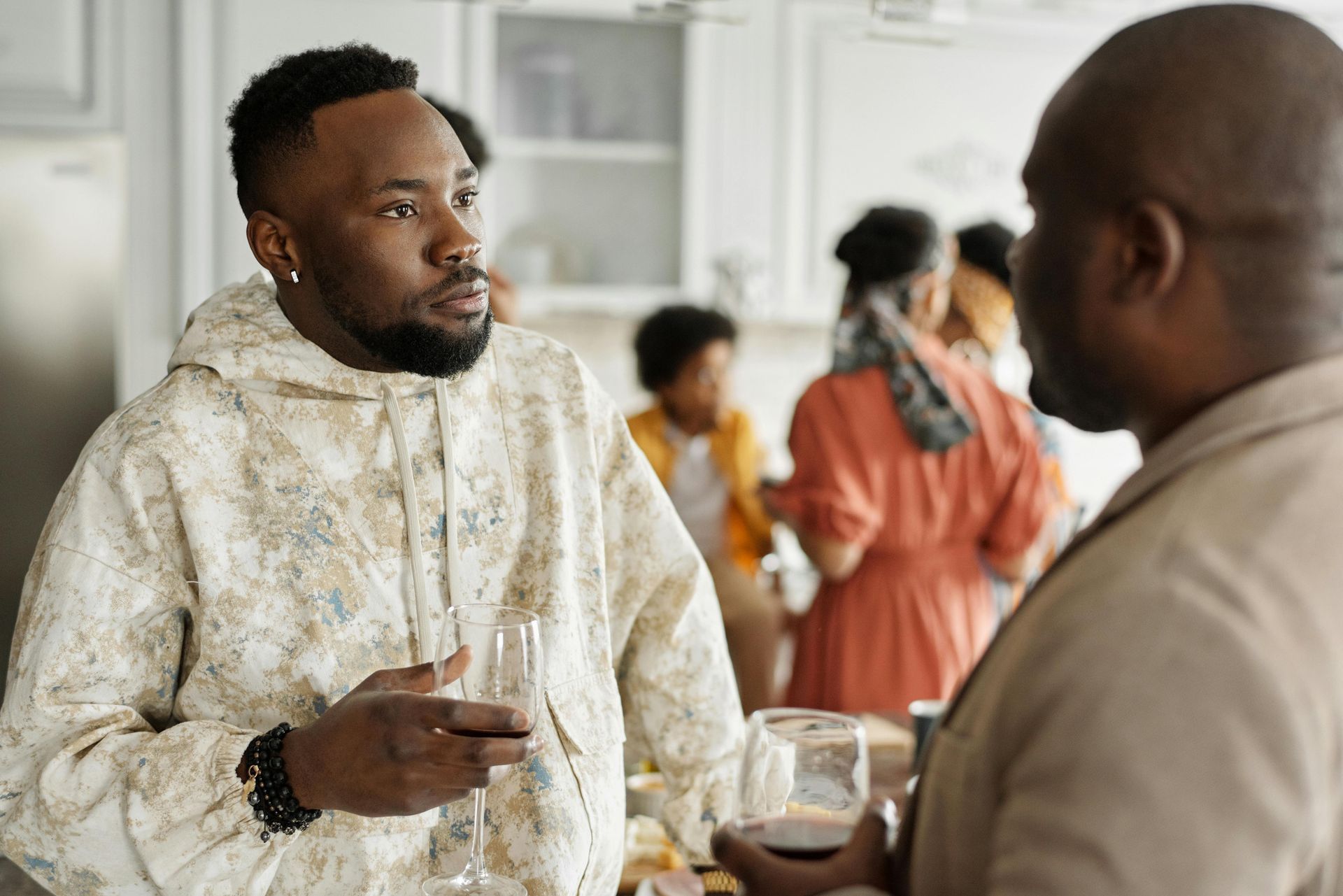 Two individuals in a kitchen hold glasses and converse, while others mingle in the background.