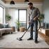 Man vacuuming a living room carpet. He wears jeans and a gray shirt. The room has a neutral color palette.