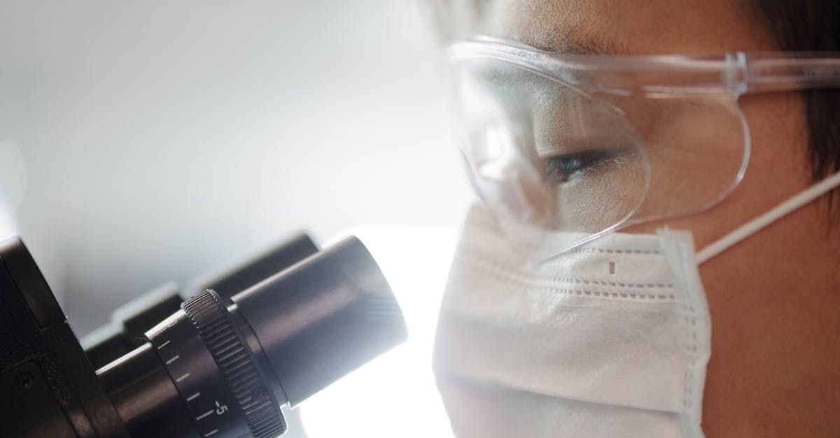 A woman wearing a mask and goggles is looking through a microscope.