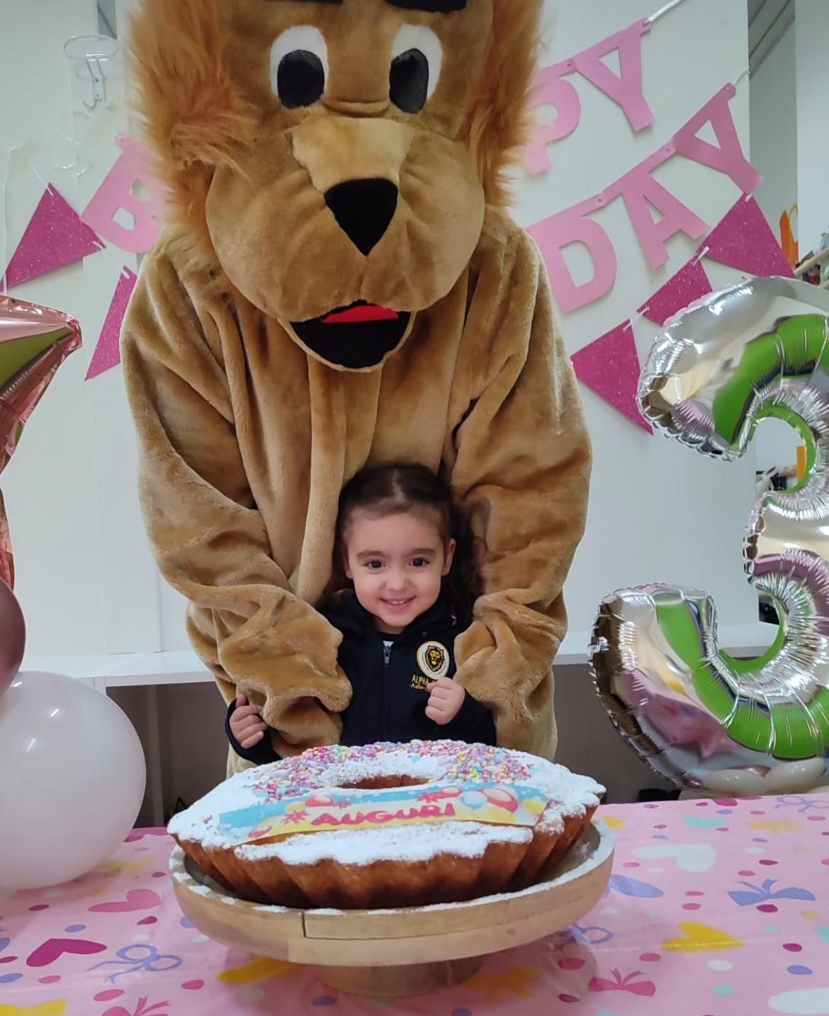 Una mascotte leone tiene in braccio una bambina davanti a una torta di compleanno