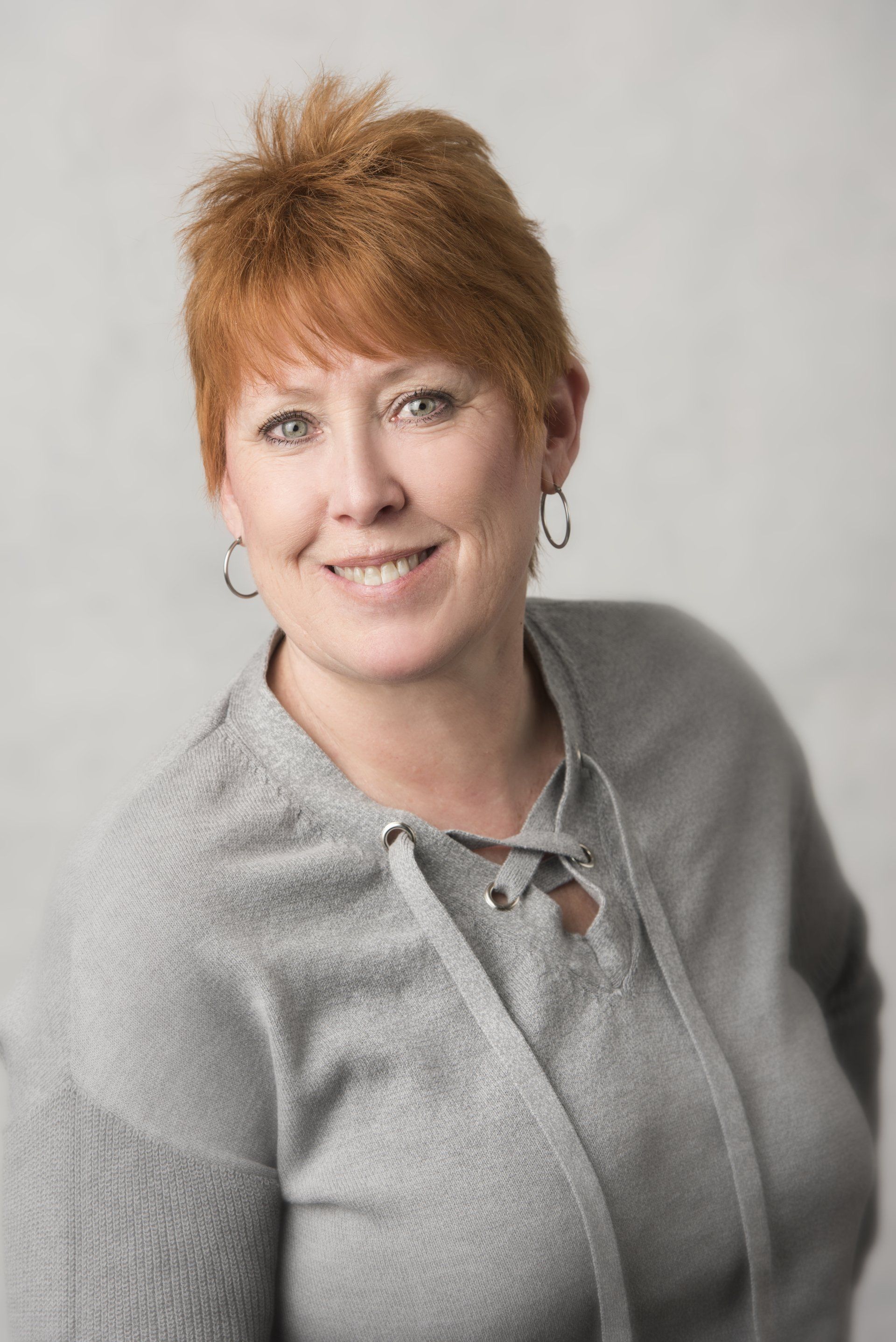 A woman wearing a grey sweater and earrings is smiling for the camera.