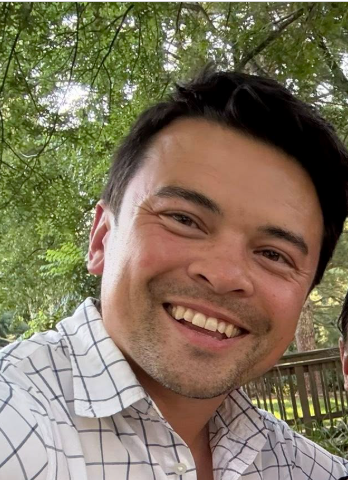 Football coach with arms crossed, looking stern on a sunny field, players in the background.