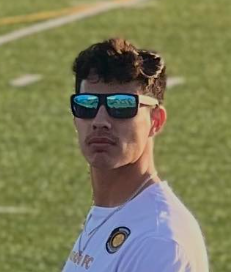 Man in green polo stands on a football field, arms crossed, stadium in background.