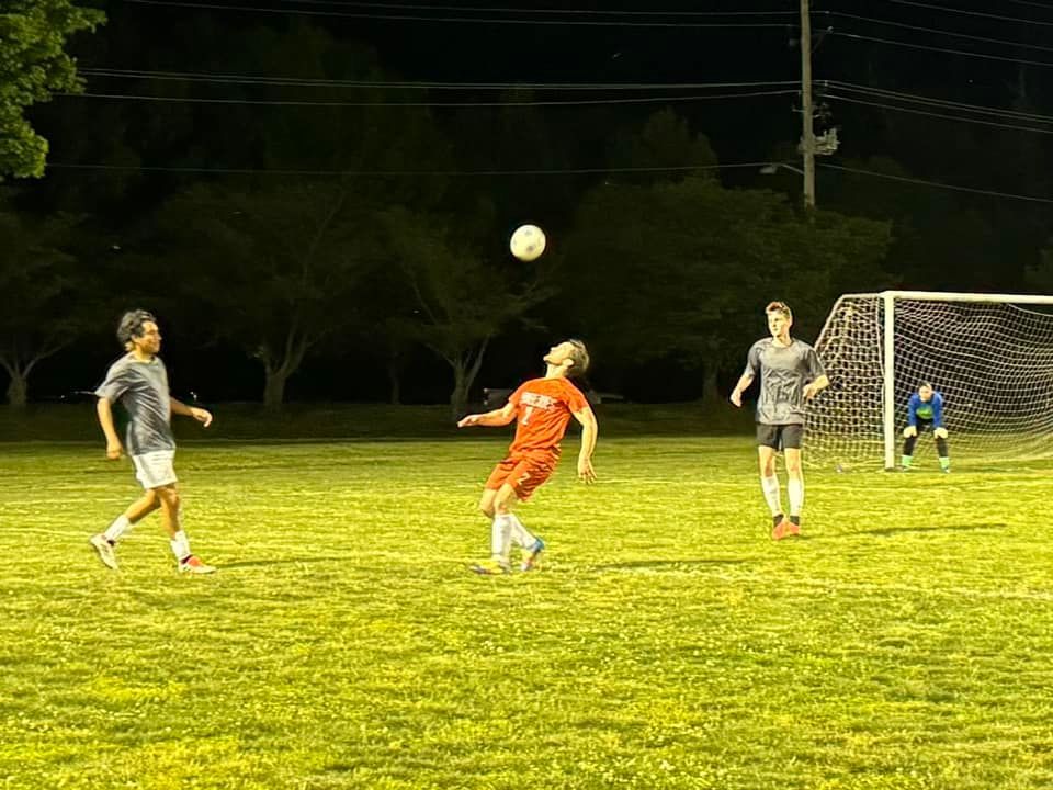 A person wearing a hat and orange shirt, holding a baseball bat on their shoulder on a baseball field.