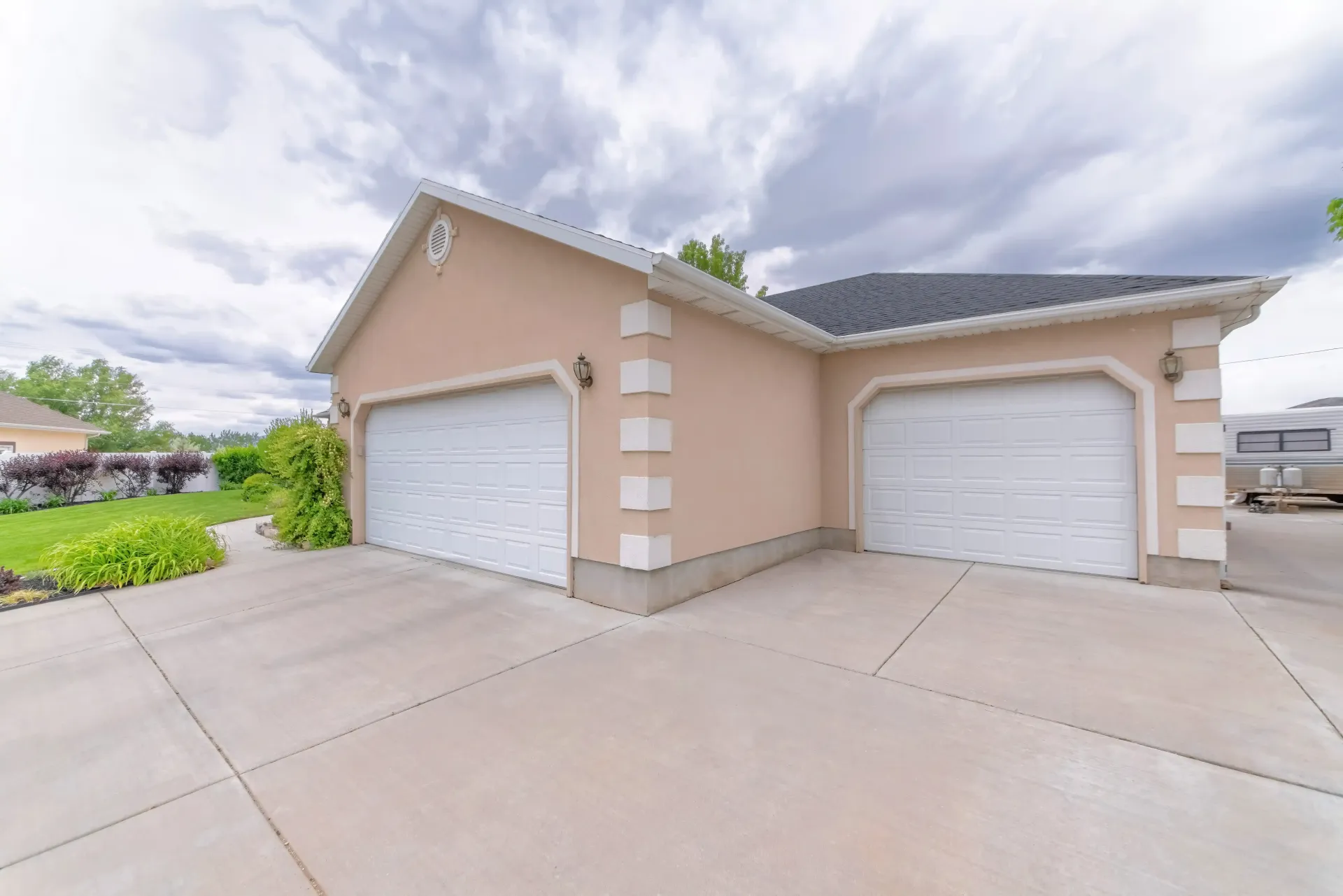 Two-car beige garage with white doors and a wide concrete driveway under a cloudy sky