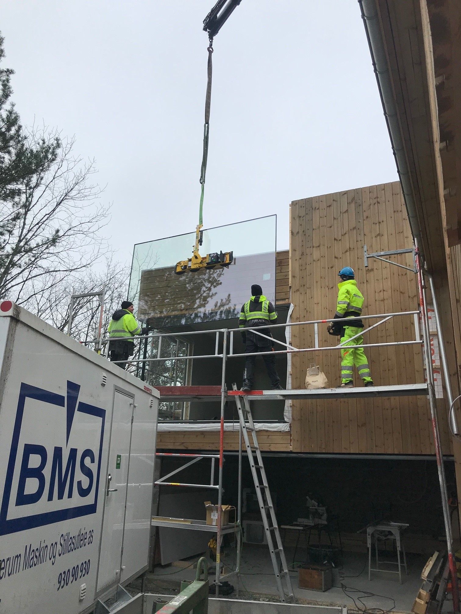 Arbeidere monterer en stor glassrute på en bygning ved hjelp av kran og stillas på en overskyet dag.