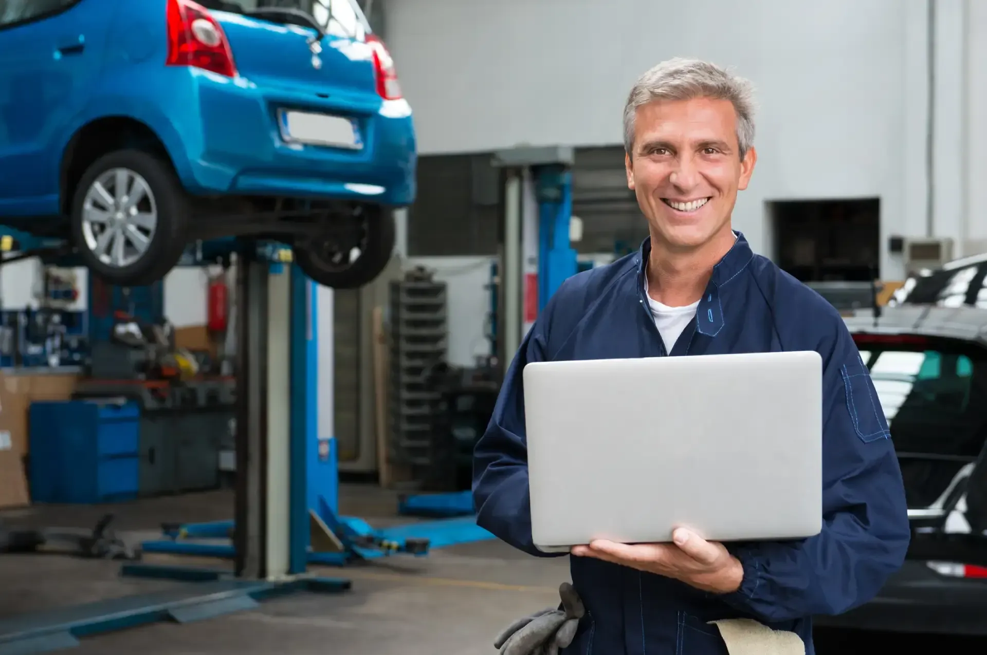 Mechanic holding a laptop in an auto repair shop, with a blue car on a lift in the background