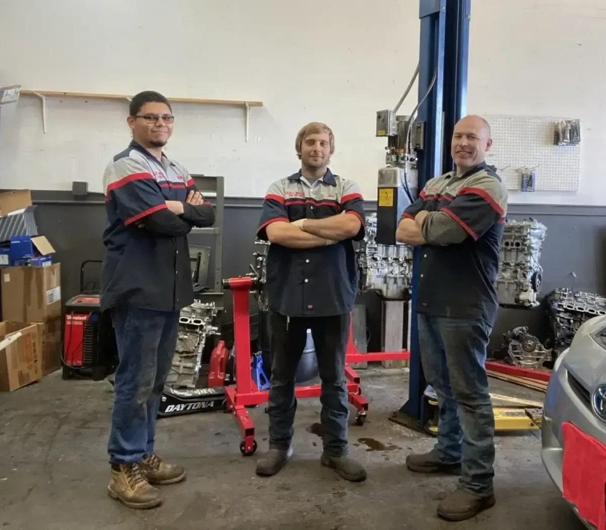 Three mechanics standing in a repair shop with arms crossed, in front of a lifted vehicle and tools.