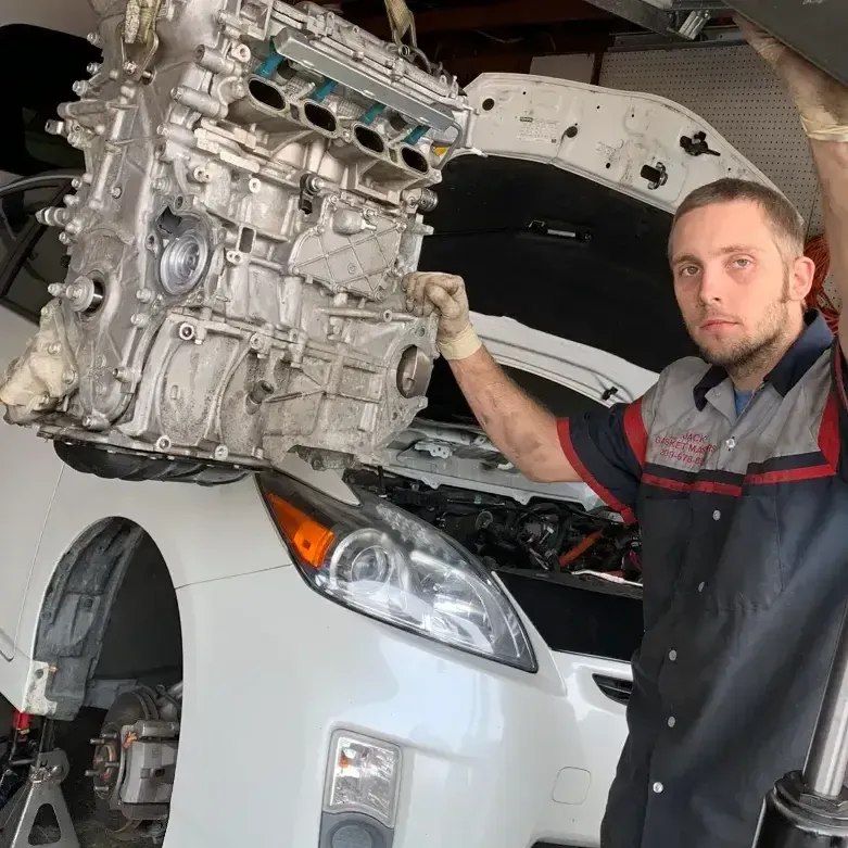 Mechanic stands beside a white car with its hood open, lifting an engine in a repair shop.
