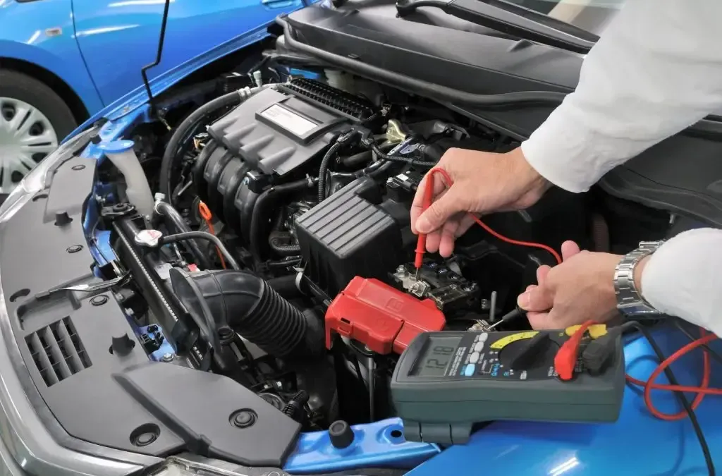 Person testing a blue car battery under the hood with jumper cables and a multimeter