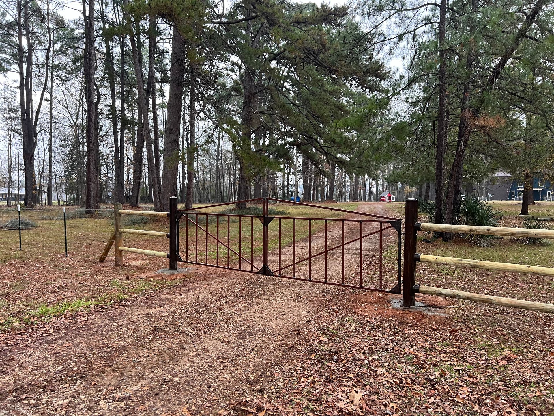 Brown metal gate on a dirt road, flanked by wooden fences, trees in background.