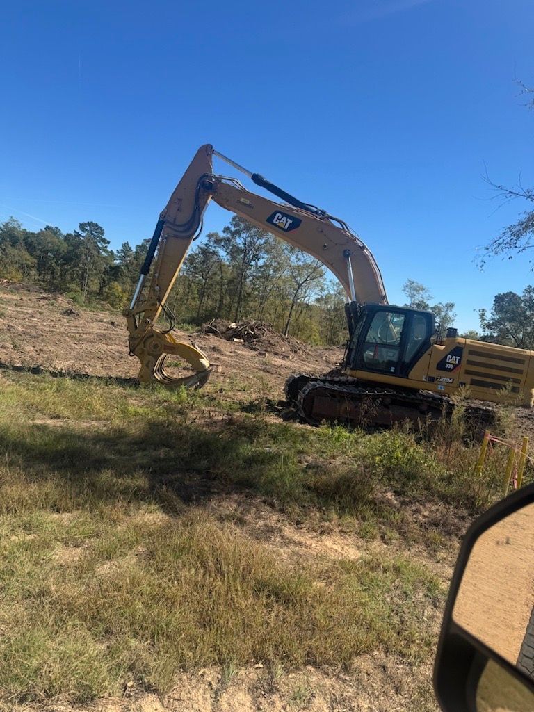 Yellow Caterpillar excavator with grapple attachment in a field clearing trees under a blue sky.