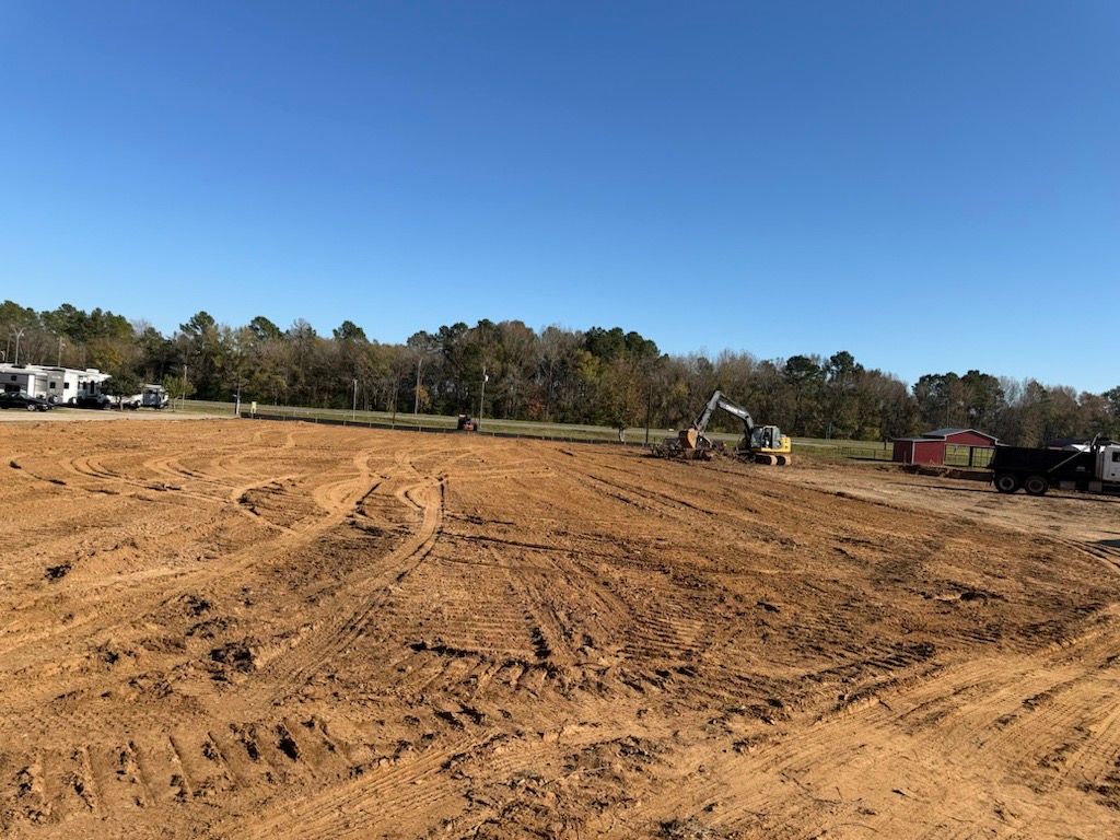 Construction site with excavator on a dirt field, trees and blue sky in the background.