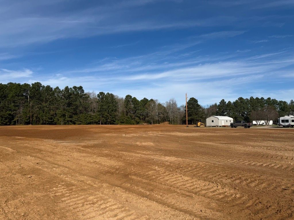 Cleared dirt field under a blue sky with trees in the background and a few buildings.