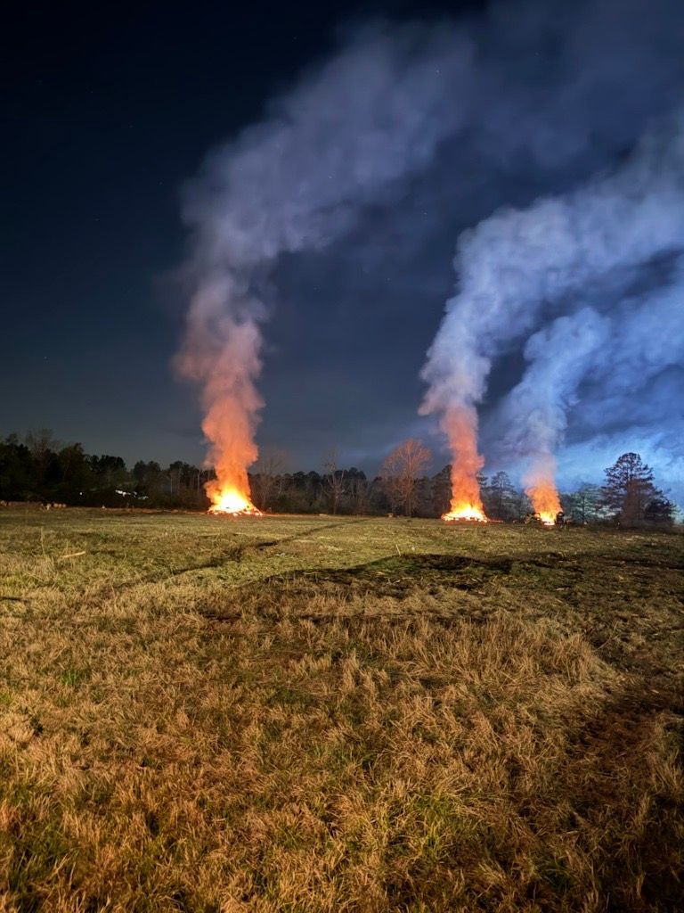 Night scene: three large fires burning in a grassy field, sending plumes of smoke into a dark sky.