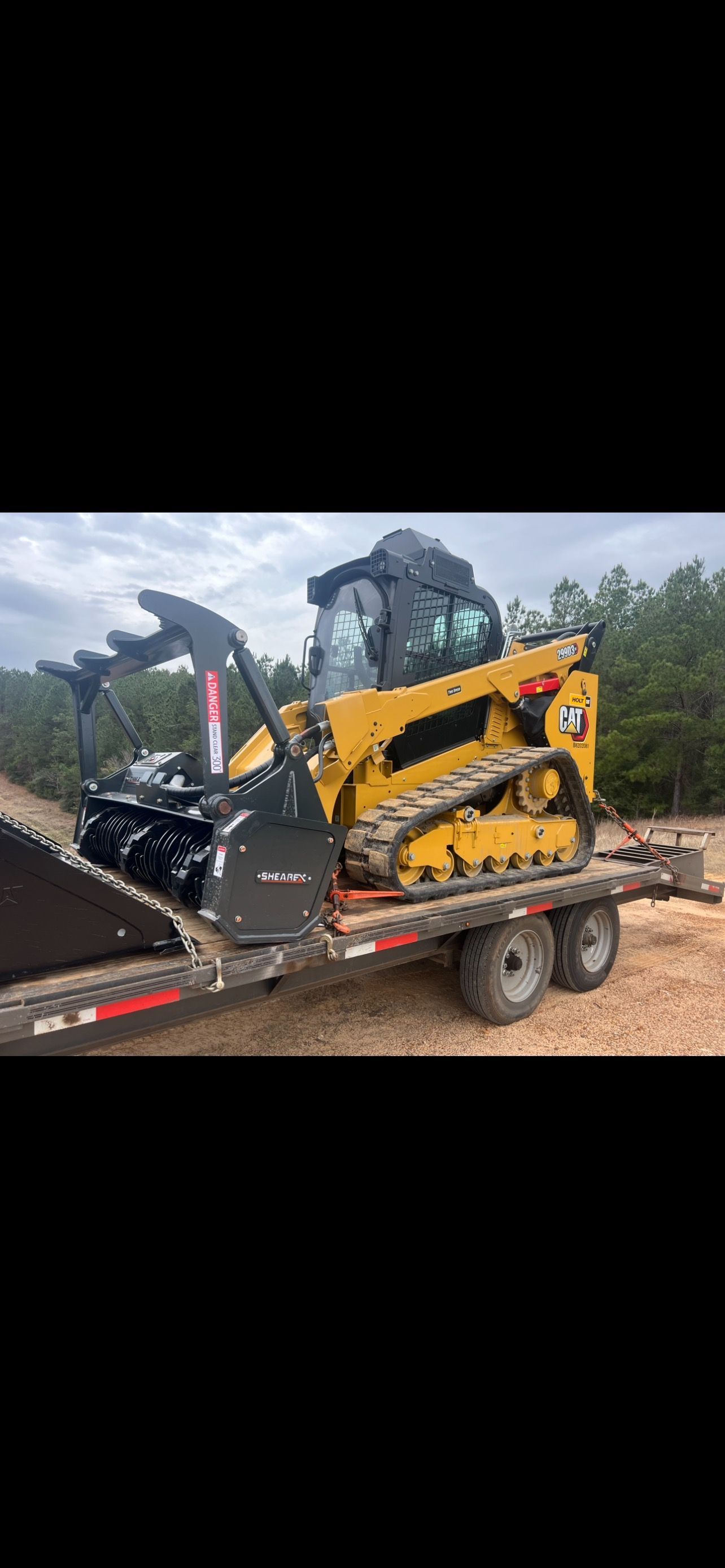 Yellow skid steer with forestry mulcher attachment on a trailer. The sky is overcast.