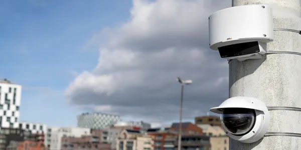 Two security cameras are mounted to a pole in a city.