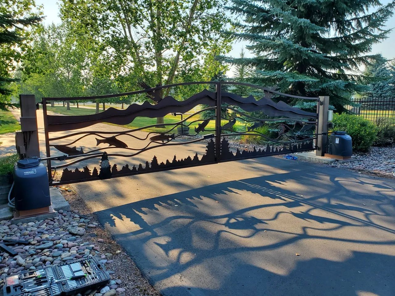 A metal gate with a silhouette of trees on it is in the middle of a driveway.