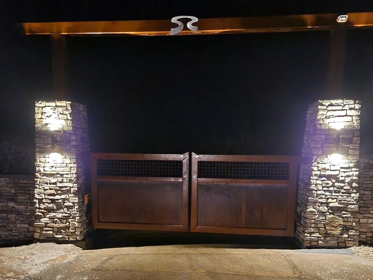 A wooden gate is surrounded by stone pillars and lights at night.