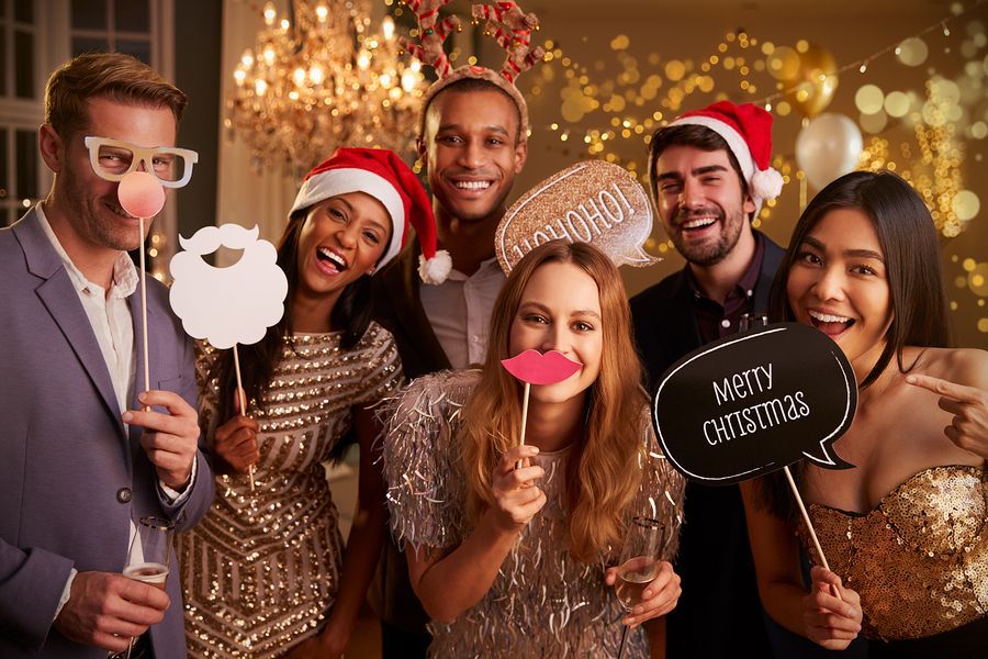 Group of friends at a party, wearing Santa hats and holding props, smiling. Festive setting with lights and decorations.