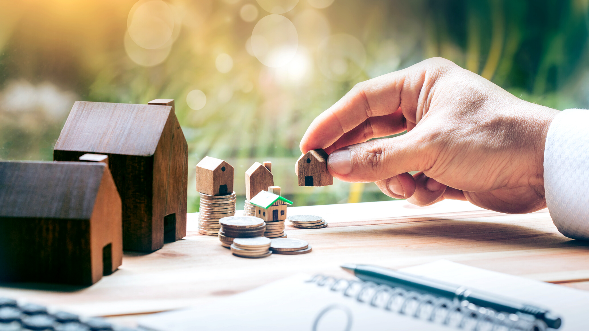 A person is holding a wooden house next to a pile of coins.