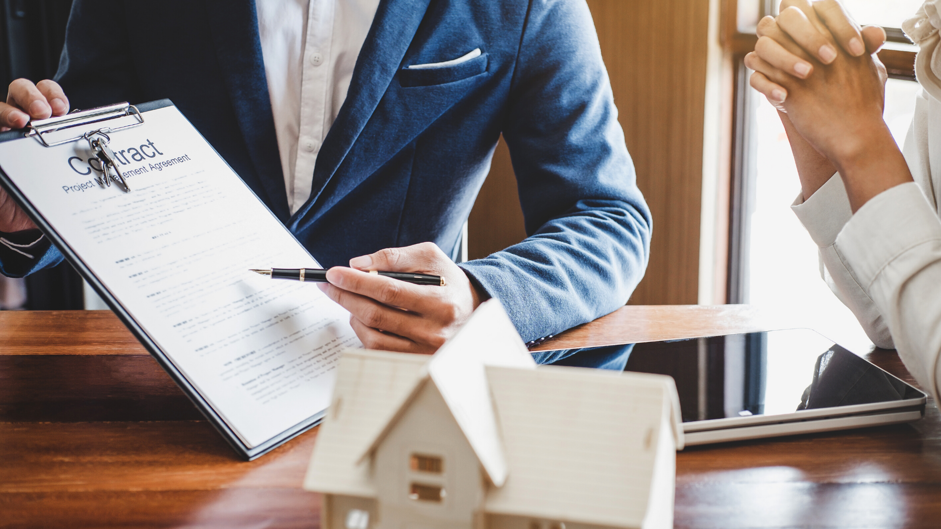 A man and a woman are sitting at a table with a model house and a clipboard.