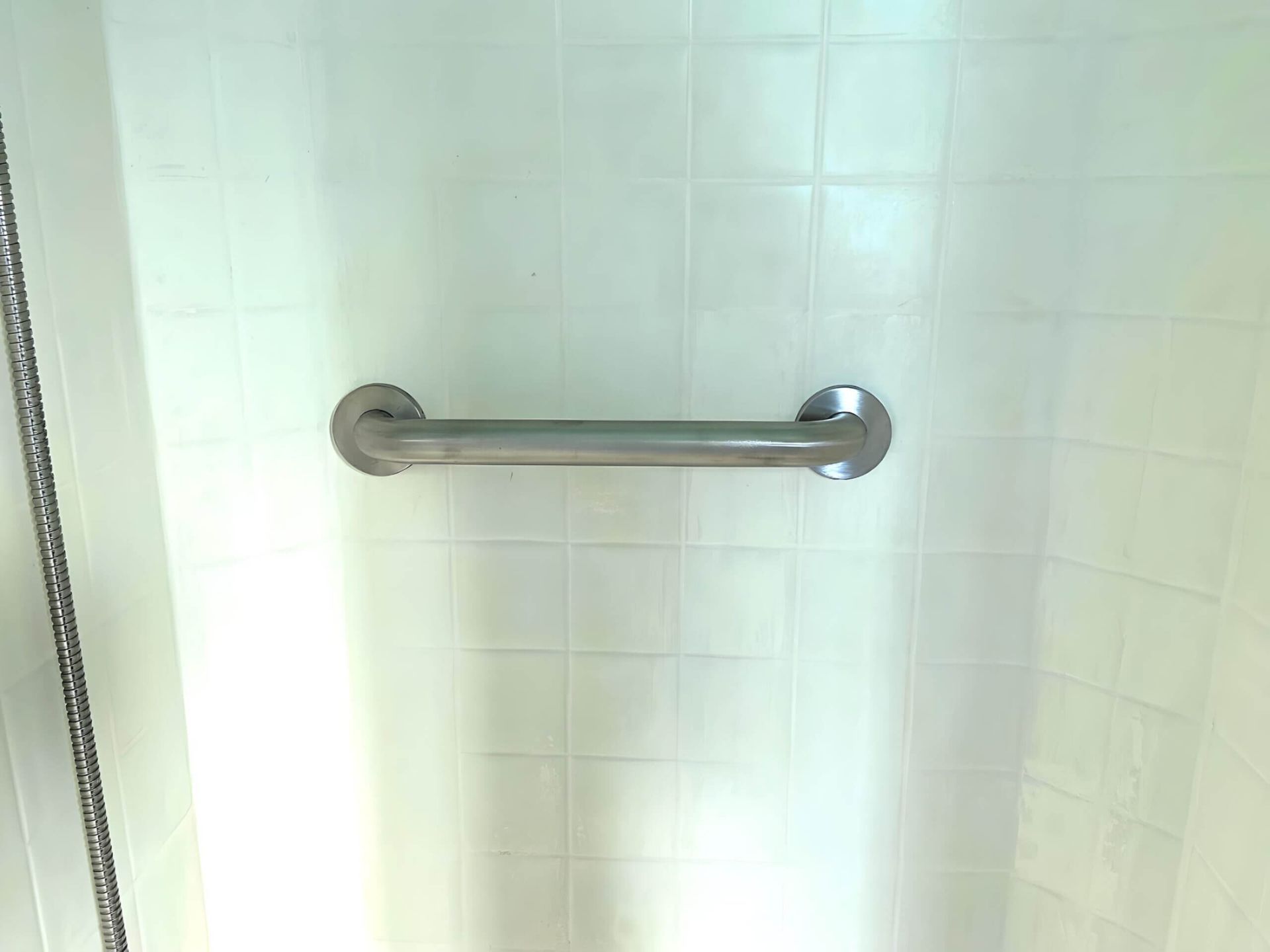 A stainless steel grab bar is hanging on a white tiled wall in a bathroom.