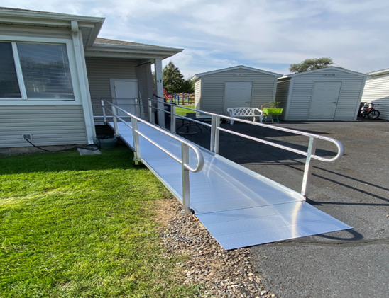 Aluminum wheelchair ramp with handrails leading from a house entrance down to a paved driveway, providing accessible entry.]