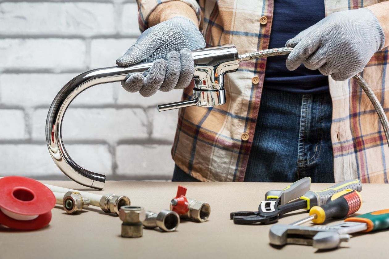 Hands in gray gloves holding a chrome faucet over a workbench with plumbing tools and parts