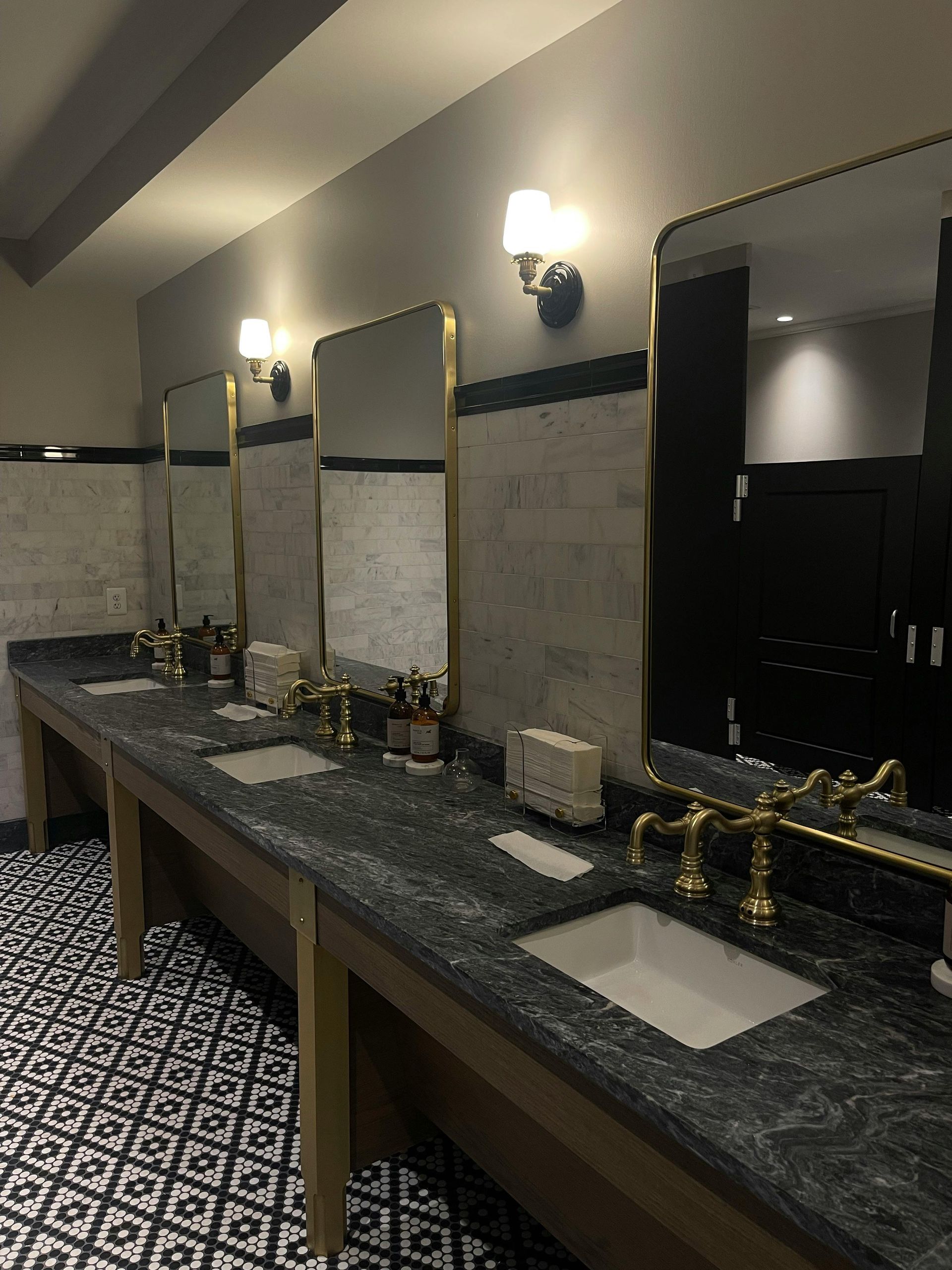 Modern restroom with a long granite countertop, mirrors, sinks, and wall sconces along patterned tile flooring