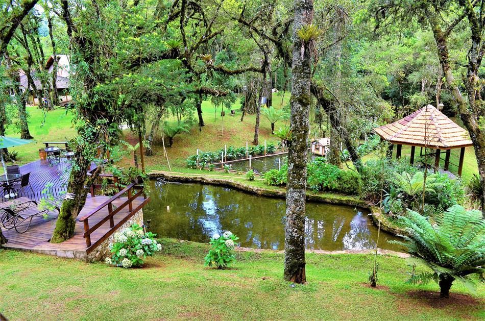 Jardim exuberante com lago, ponte de madeira, degraus de pedra e um gazebo de palha rodeado por árvores.