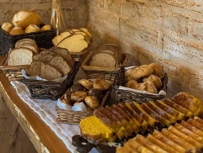 Cesta com pães e doces variados sobre uma mesa coberta com uma toalha, junto a uma parede de tijolos.