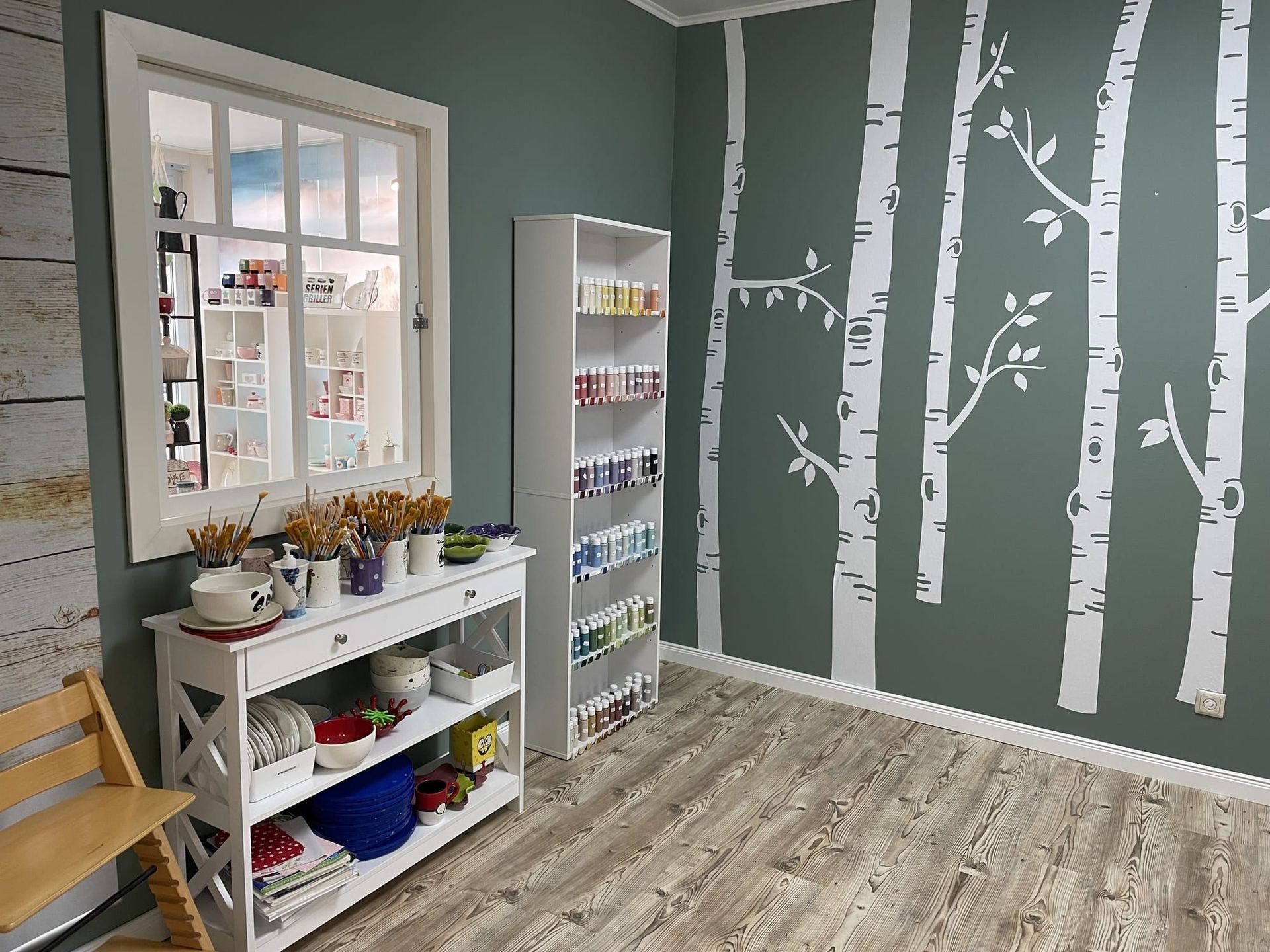 Ein Raum mit einem Regal und einem Fenster, an dessen Wand Bäume gemalt sind.