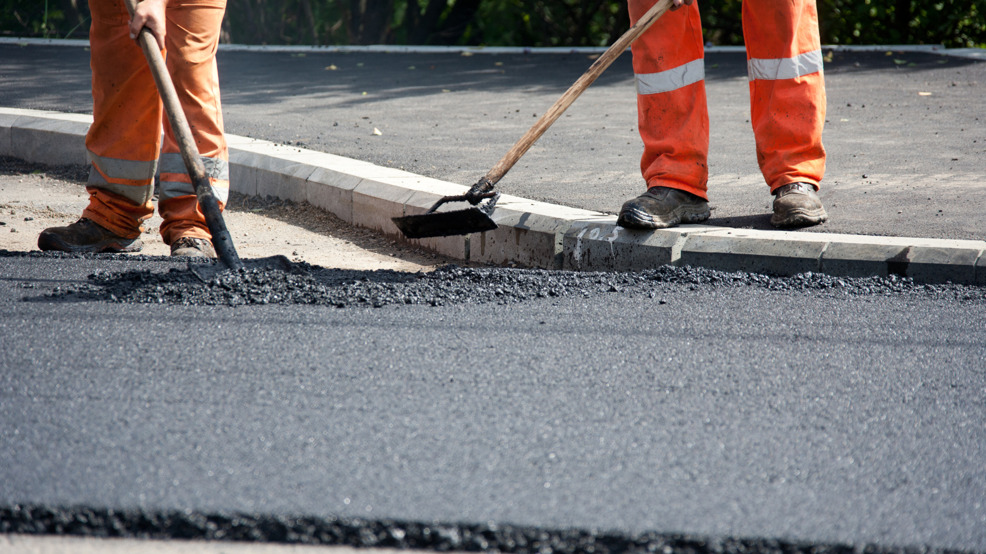 Two road workers in bright orange uniforms use rakes to spread fresh hot asphalt on a street.