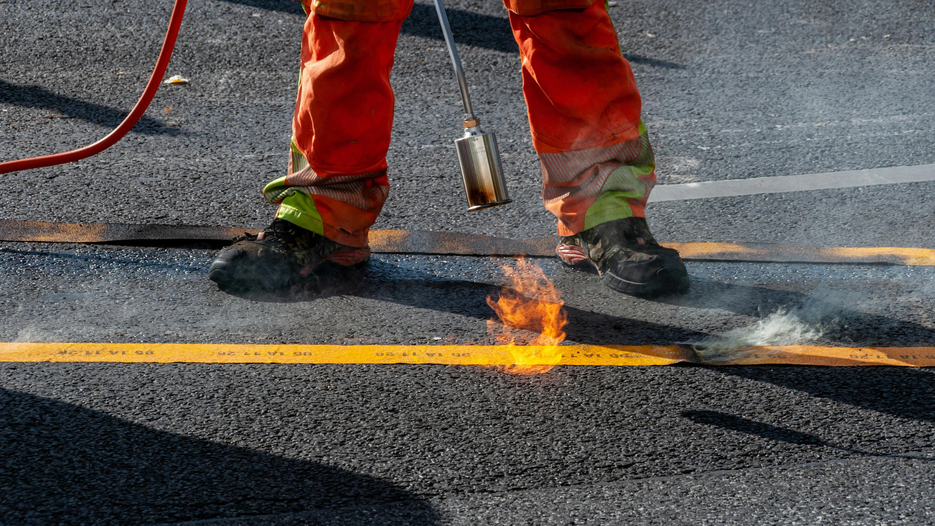 A worker in orange high-visibility trousers uses a torch to melt yellow thermoplastic pavement markings onto asphalt.
