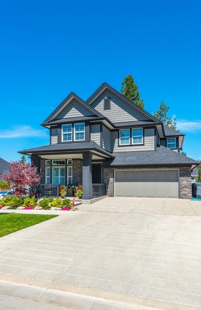 Two-story gray house with stone facade, two-car garage, and paved driveway under a blue sky.
